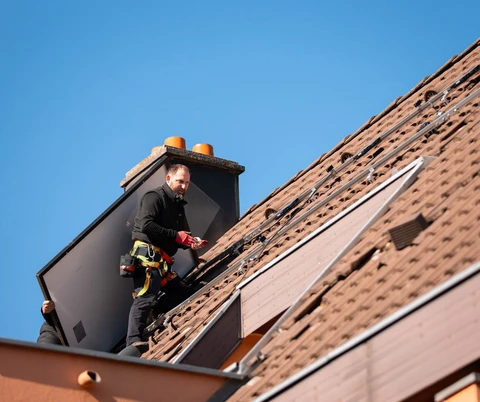 Technicien en combinaison noire et harnais de sécurité rouge installe des panneaux photovoltaïques sur toiture en tuiles terre cuite