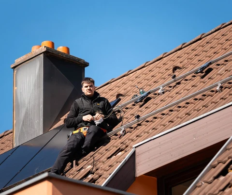Technicien en combinaison noire équipé d'un harnais de sécurité orange, assis sur une toiture en tuiles romanes rouges, installant des panneaux photovoltaïques