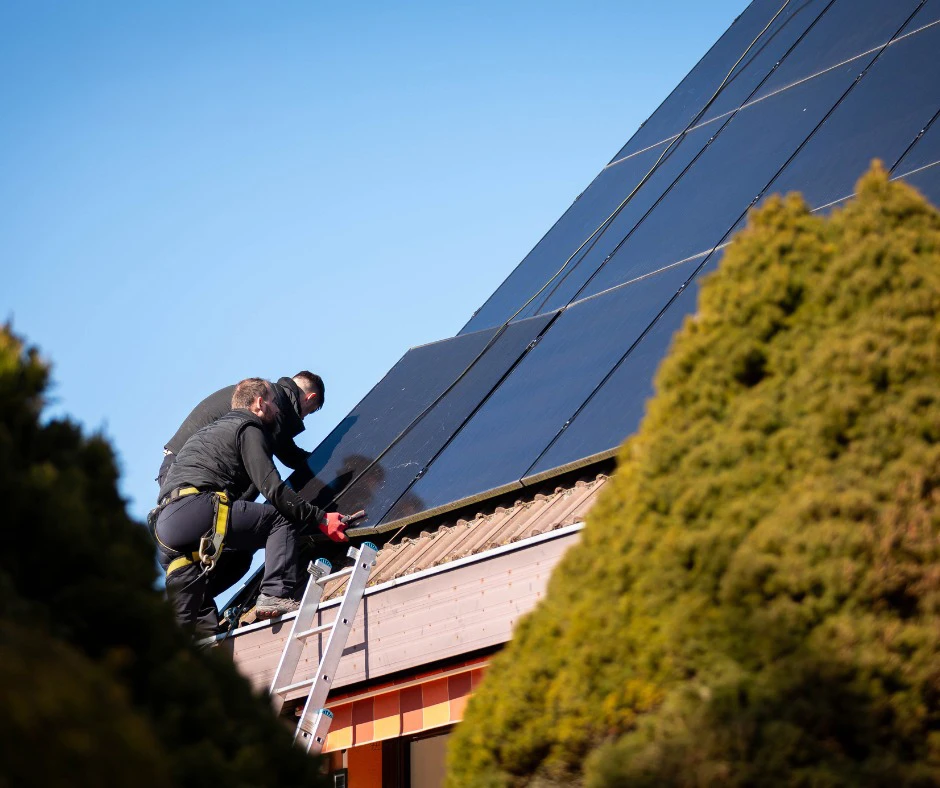 Deux techniciens en combinaison noire installent des panneaux solaires photovoltaïques sur une toiture en pente, depuis une échelle en aluminium
