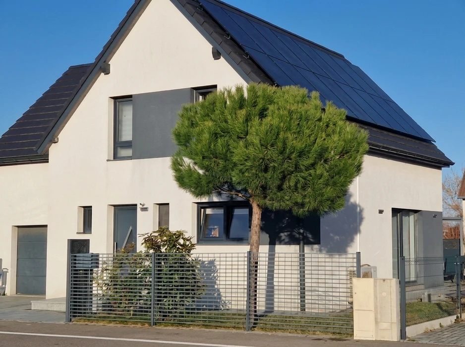 Maison individuelle moderne, façade blanche avec bardage gris anthracite, toiture en tuiles noires équipée de panneaux photovoltaïques full-black